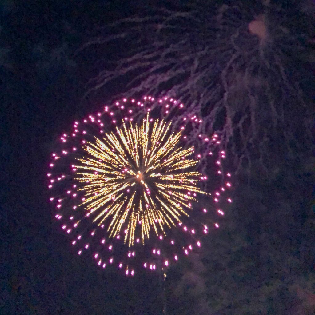 yellow and purple fireworks bloom against a night sky, with smoke trails from previous bursts